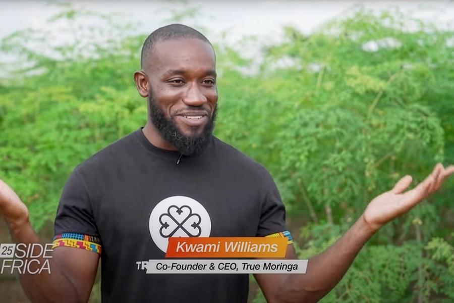 Kwami Williams, Co-Founder and CEO of True Moringa, speaks on CNN’s Inside Africa program. He smiles with arms raised slightly, standing outdoors in front of lush green moringa plants.